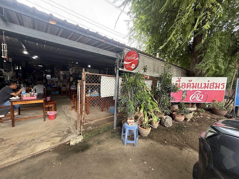 ทางเข้าร้านจากริมถนน มีป้ายชื่อร้าน ไม้กระถาง และโต๊ะใต้หลังคา