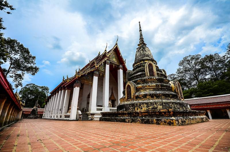 อุโบสถเก่าเสาสีขาวกับเจดีย์ทรงระฆังบนลานอิฐ ใต้ท้องฟ้ามีเมฆ